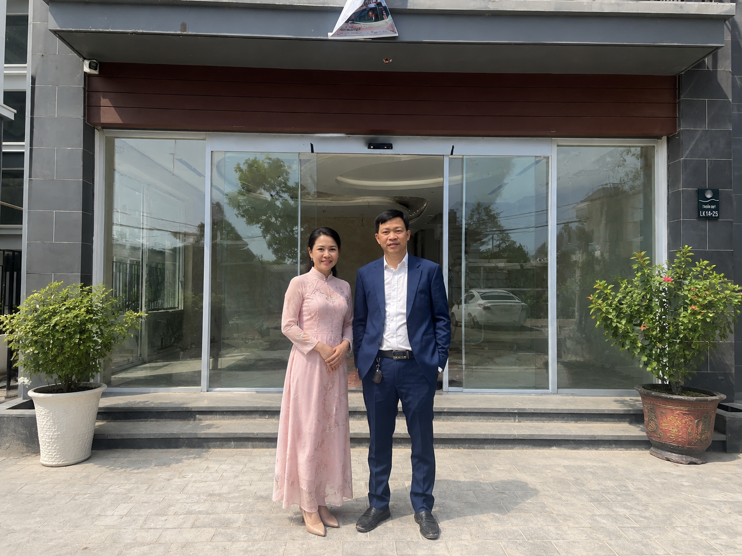 Cặp đôi mặc áo dài hồng và vest xanh đứng trước cửa kính tòa nhà văn phòng hiện đại